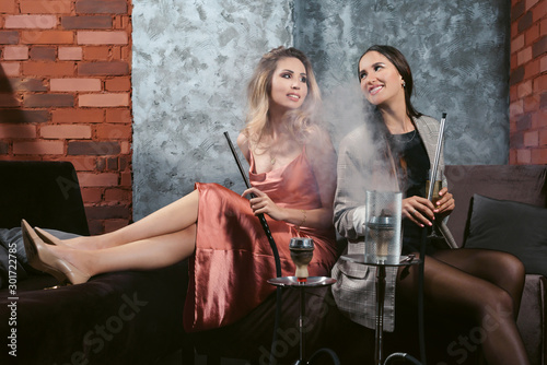 Girls party in hookah lounge. Group of two young women smoking shisha in cafe or bar, making smoke clouds, having fun, smiling. Relax concept. Friendship