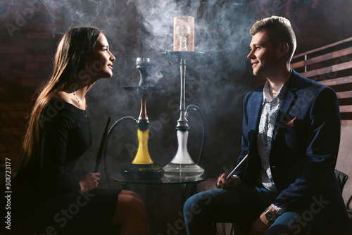 Beautiful couple woman and man in backlight in love smoking hookah and drinks cocktails in the bar