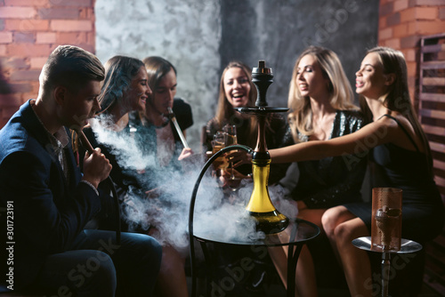 Friends party in hookah lounge. Group of people women and men smoking shisha in cafe or bar, making smoke clouds, having fun, smiling. Relax concept. Friendship photo
