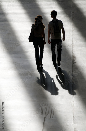 Silhouette of unrecognizable couple walking on a modern concrete walkway in dramatic shadows alternating with shafts of sunlight