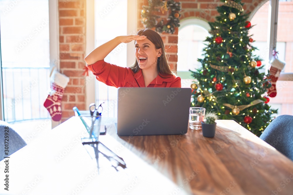 Beautiful woman sitting at the table working with laptop at home around christmas tree very happy and smiling looking far away with hand over head. Searching concept.