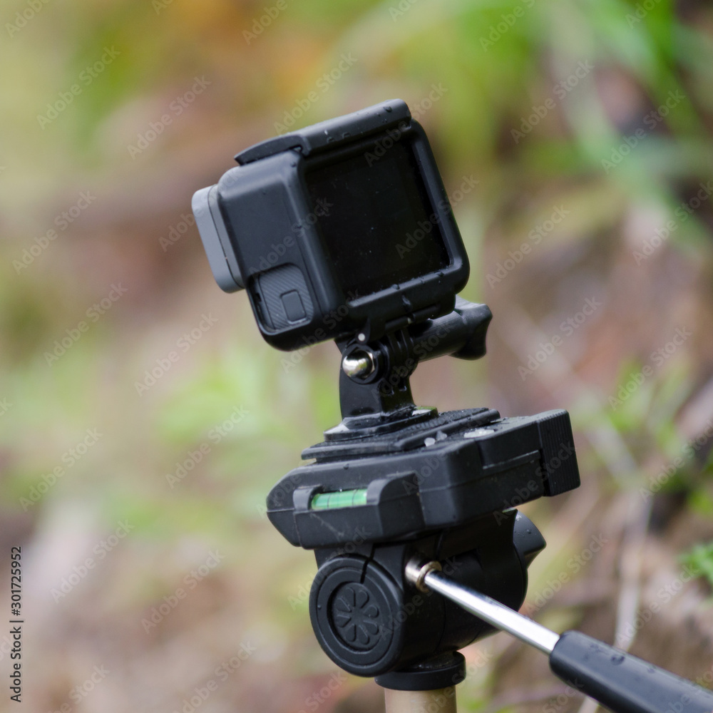 Action camera stands on a small tripod with a water level, recording video, among the grass and forest.