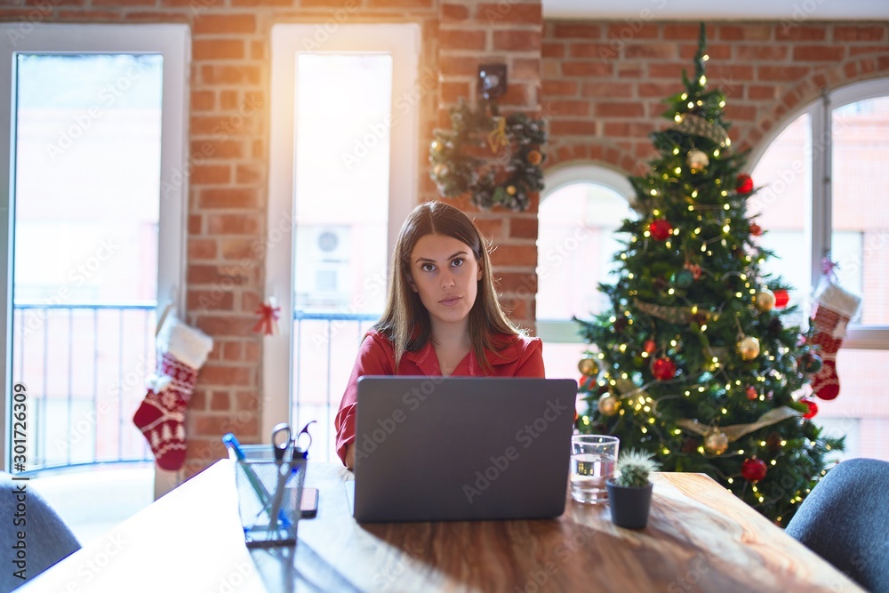 Beautiful woman sitting at the table working with laptop at home around christmas tree Relaxed with serious expression on face. Simple and natural looking at the camera. Beautiful woman sitting at the table working with laptop at home around christmas tree Relaxed with serious expression on face. Simple and natural looking at the camera.