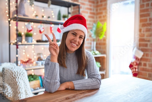 Young beautiful woman wearing christmas hat sitting at the table at home smiling with happy face winking at the camera doing victory sign. Number two.