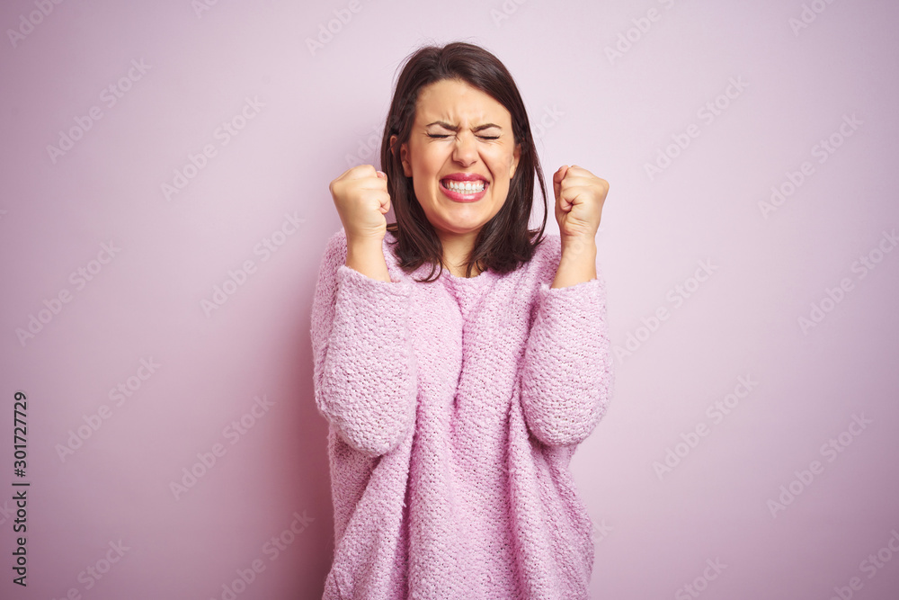 Young beautiful brunette woman wearing a sweater over pink isolated background excited for success with arms raised and eyes closed celebrating victory smiling. Winner concept. Young beautiful brunette woman wearing a sweater over pink isolated background excited for success with arms raised and eyes closed celebrating victory smiling. Winner concept.