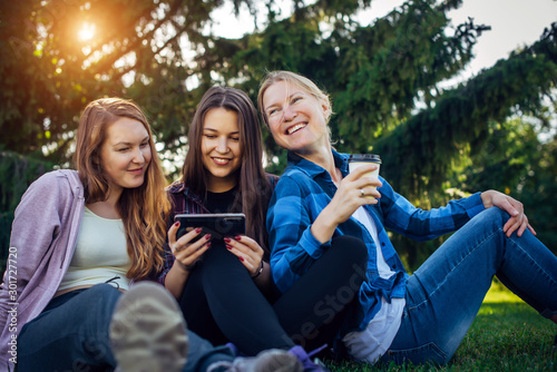 Happy young girls have fun laughing sitting on green grass in the park. Students in between lectures. Blonde, brunette and red-hair girl communicate, look at smartphone, concept of female friendship.
