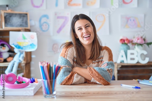 Young beautiful teacher woman wearing sweater and glasses sitting on desk at kindergarten happy face smiling with crossed arms looking at the camera. Positive person.
