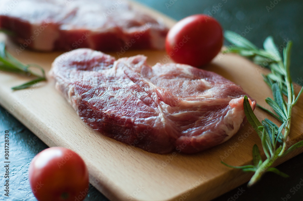 Raw pork steak, rosemary and tomato on a cutting board, on a dark gray concrete background, close up, in dark tonality