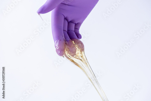 Woman's hand in a glove holds yellow sugar paste or wax for depilation close up on a white background