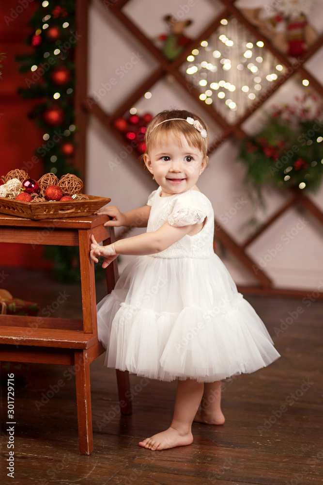 New Year and Christmas celebration concept. Pretty little girl in white dress playing and being happy about christmas tree and lights. Winter holidays.