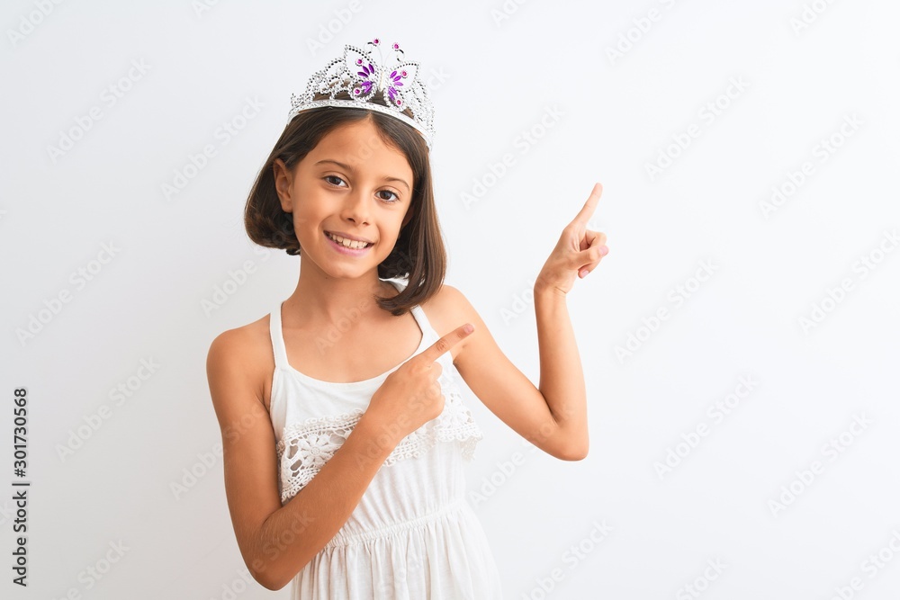 Beautiful child girl wearing princess crown standing over isolated white background smiling and looking at the camera pointing with two hands and fingers to the side. Beautiful child girl wearing princess crown standing over isolated white background smiling and looking at the camera pointing with two hands and fingers to the side.