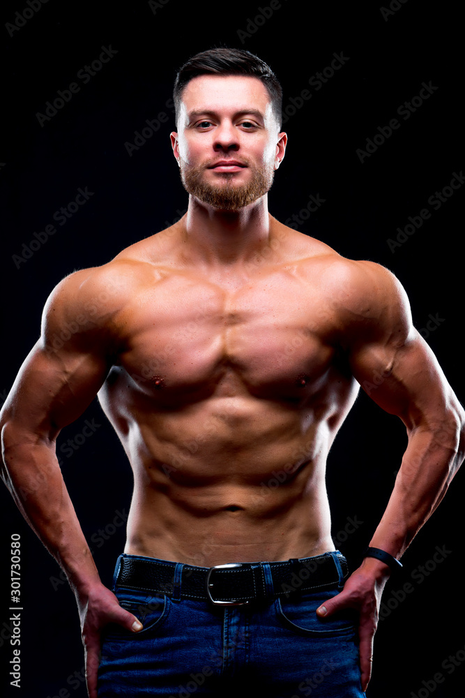 Beautiful young man with perfect body posing on dark background. Strong hands on waist. Studio photoshoot.