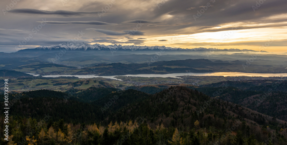 Dark landscape of the mountains at sunset. Autumn view of the mountain range, evening fog at their foothills and town on the lake in the valley.