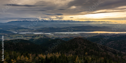 Dark landscape of the mountains at sunset. Autumn view of the mountain range, evening fog at their foothills and town on the lake in the valley.