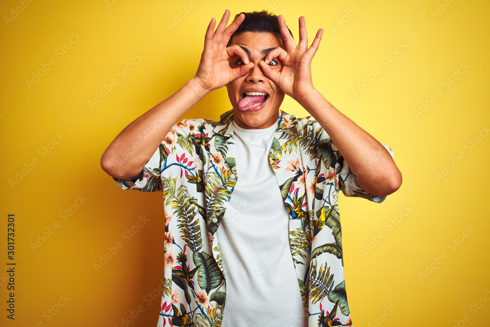 Young brazilian man on vacation wearing summer floral shirt over isolated yellow background doing ok gesture like binoculars sticking tongue out, eyes looking through fingers. Crazy expression. Young brazilian man on vacation wearing summer floral shirt over isolated yellow background doing ok gesture like binoculars sticking tongue out, eyes looking through fingers. Crazy expression.