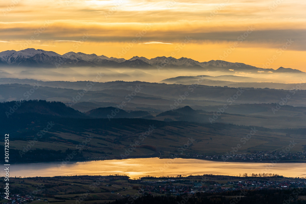 Beautiful fairytale landscape of the mountains at sunset. Autumn view of the mountain range with peaks in the snow, evening fog over the lowlands and the lake where the towns were located.