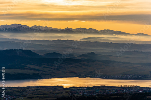 Fototapeta Naklejka Na Ścianę i Meble -  Beautiful fairytale landscape of the mountains at sunset. Autumn view of the mountain range with peaks in the snow, evening fog over the lowlands and the lake where the towns were located.