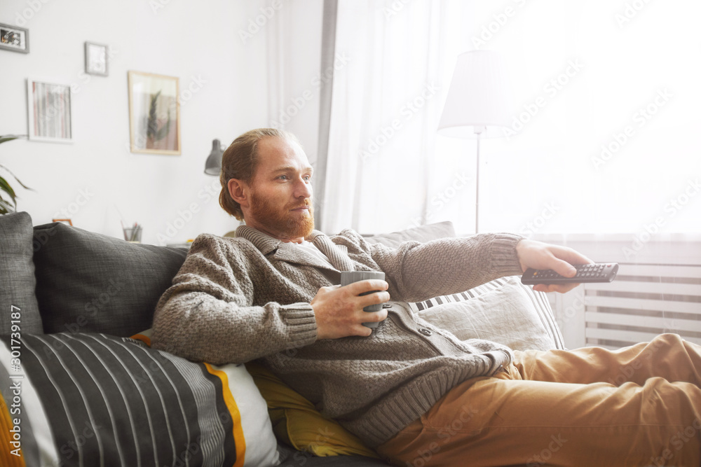 Side view portrait of middle-aged bearded man watching TV at home and switching channels via remote control while relaxing on comfortable couch