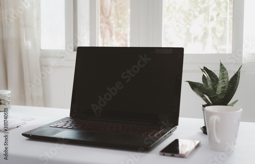 Black laptop is placed on a white desk with in the office with windows and ornamental plants. Business objects concept