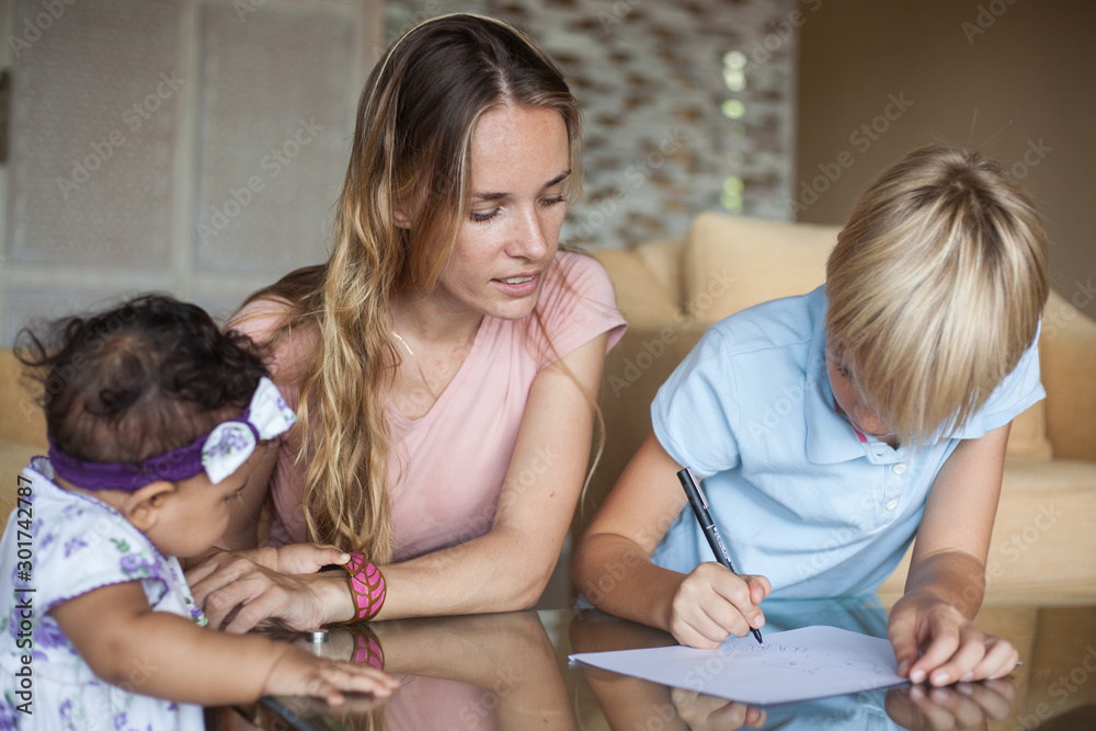 young attractive mother with her 10 y.o son and toddler daughter in the house at the table draw on a white sheet of paper. Multiracial family. young attractive mother with her 10 y.o son and toddler daughter in the house at the table draw on a white sheet of paper. Multiracial family.