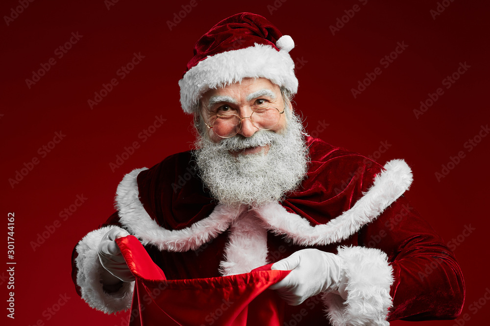 Waist up portrait of smiling Santa Claus holding sack with Christmas presents while standing over red background, copy space Waist up portrait of smiling Santa Claus holding sack with Christmas presents while standing over red background, copy space