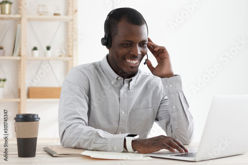 Dark-skinned guy from technical support wearing headset with microphone, talking to clients and checking data on laptop photo