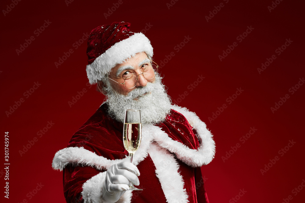 Waist up portrait of smiling Santa Claus holding champagne glass while standing over red background, copy space Waist up portrait of smiling Santa Claus holding champagne glass while standing over red background, copy space