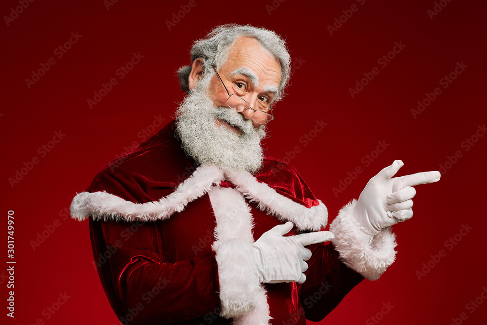 Waist up portrait of smiling Santa Claus looking at camera and pointing to side while standing against red background, copy space