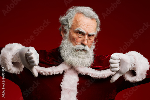 Waist up portrait of frowning Santa Claus showing thumbs down while posing against red background in studio photo