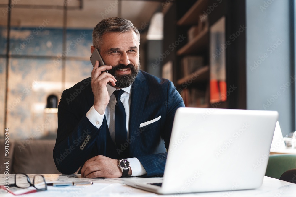 Portrait of a nice smiling man with beard, he is sitting at table, talking on the phone with his laptop Portrait of a nice smiling man with beard, he is sitting at table, talking on the phone with his laptop