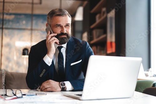Portrait of a nice smiling man with beard, he is sitting at table, talking on the phone with his laptop