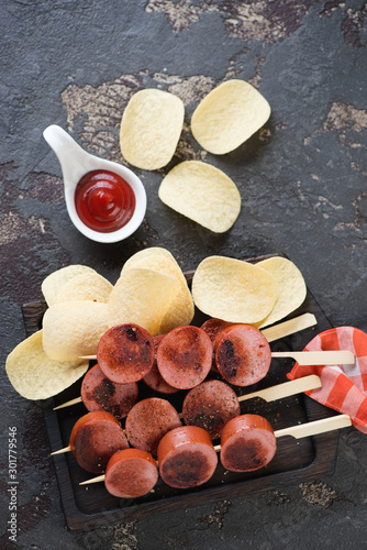 Brochettes with slices of roasted sausages and potato chips, vertical shot on a brown stone background, flatlay