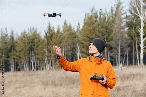 A man in an orange jacket  with a backpack and a hat  controls a quadcopter drone outdoors in autumn. The guy holds out his hand to the drone.