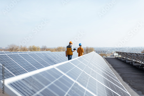 View on the rooftop solar power plant with two engineers walking and examining photovoltaic panels. Concept of alternative energy and its service photo