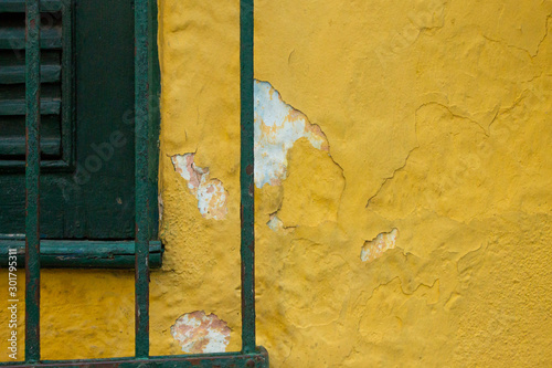 A wooden green window with a green fence in front and a yellow wall with some peeling white parts appearing
