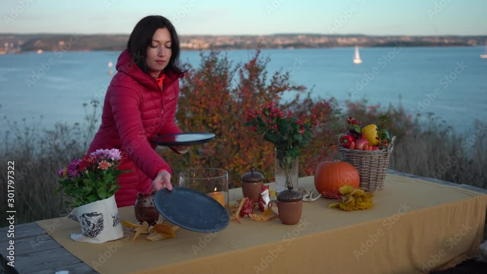 Young beautiful woman in red jacket is decorating autumn evening picnic table with seaside on blurry background Young beautiful woman in red jacket is decorating autumn evening picnic table with seaside on blurry background