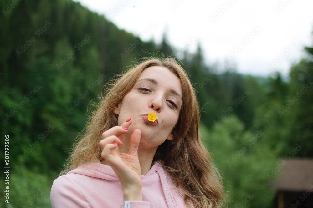 Outdoors portrait of young calm blonde woman in pink clothes with little yellow flower in her mouth on the hill with green forest background during spring or early autumn in the mountains Outdoors portrait of young calm blonde woman in pink clothes with little yellow flower in her mouth on the hill with green forest background during spring or early autumn in the mountains