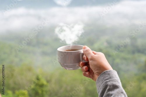 Hot coffee cup with heart shape steam in hand of woman, blurred landscape of forest in background