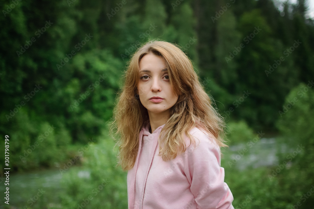 Serious young blonde woman in pink clothes with long hair is looking at the camera with anxiety on the hill with green forest background during spring or early autumn in the mountains. Serious young blonde woman in pink clothes with long hair is looking at the camera with anxiety on the hill with green forest background during spring or early autumn in the mountains.