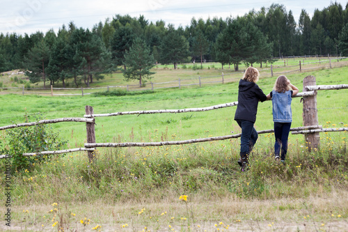 Adult woman and teenage girl enjoying nature in countryside, standing close to farm pasture, rear view