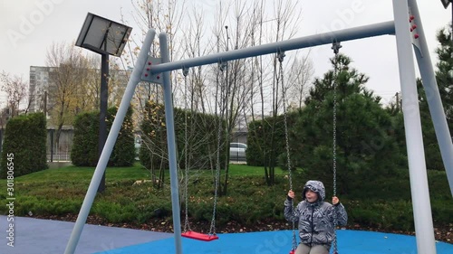 Cheerful European boy swinging on a swing. A joyful child swings on a swing in the Playground.  Happy child swinging very high on a swing in the street. Fall season. Cloudy weather. photo