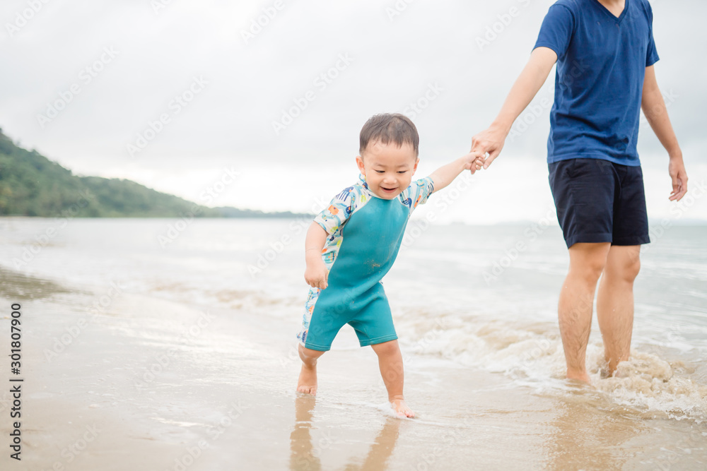 Asian toddler boy walking to the sea with his father.Dad holding hand his son for swim and play at the beach in Thailand.