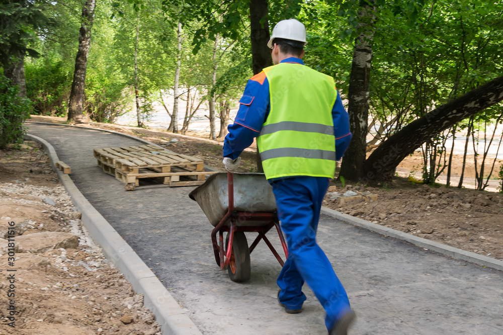 builder in workwear and helmet carries an empty cart on a new road, rear view builder in workwear and helmet carries an empty cart on a new road, rear view