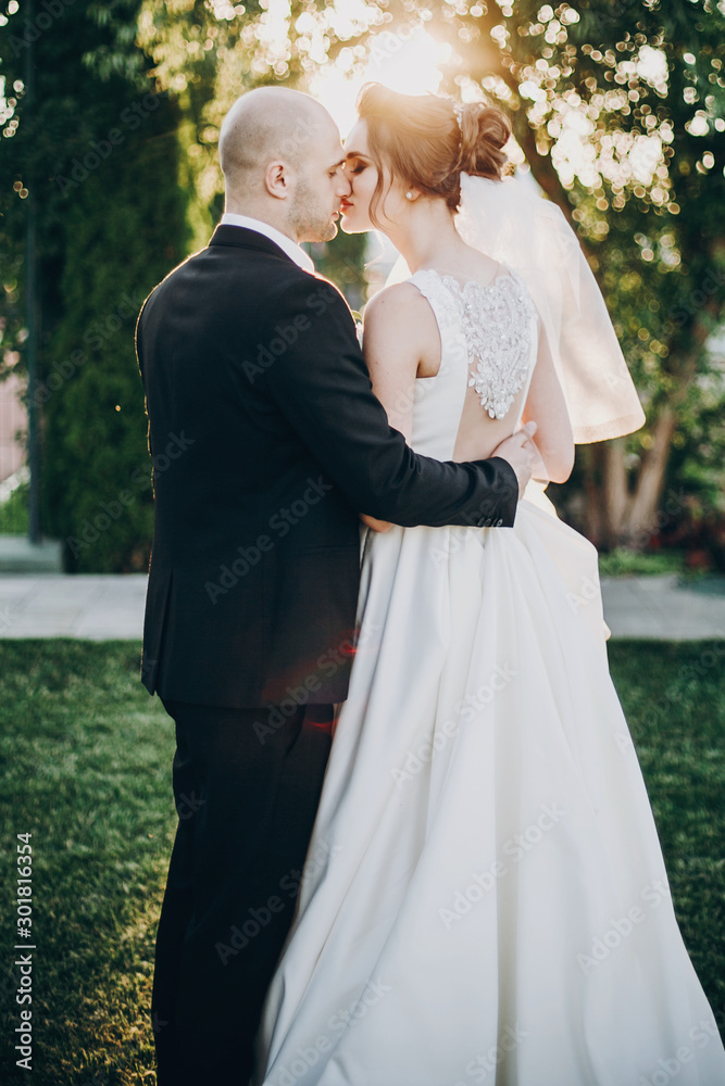 Stylish bride and groom gently kissing in sunset light. Beautiful bride with modern bouquet embracing with stylish groom in summer park. Sensual romantic moment