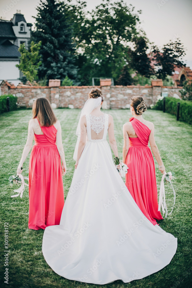 Fototapeta premium Gorgeous bride walking with bridesmaids in pink dresses, holding stylish bouquets in warm evening light in summer park. Beautiful happy bride with maid of honor posing together
