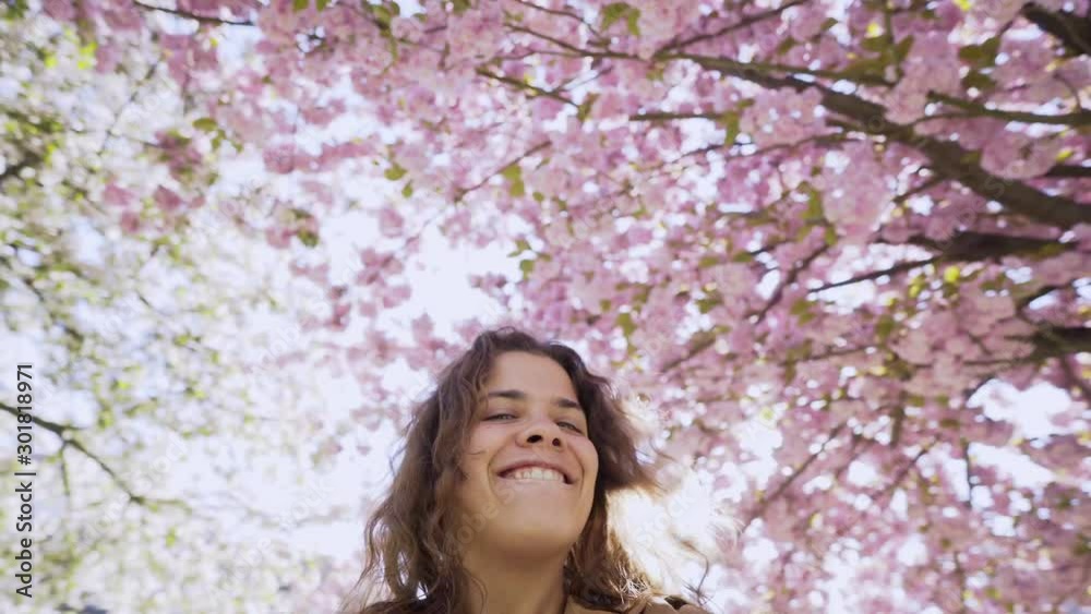 Young girl taking selfie in blooming garden at backlit. First perspective playful woman recording himself on video, looking at camera while moving and turning under blossom cherry tree in sunny day Young girl taking selfie in blooming garden at backlit. First perspective playful woman recording himself on video, looking at camera while moving and turning under blossom cherry tree in sunny day