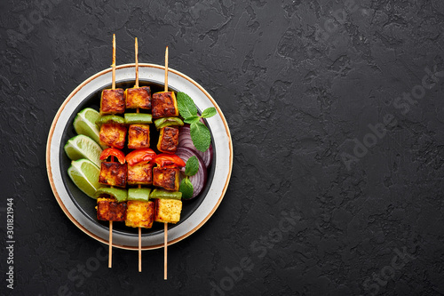 Paneer Tikka at skewers in black bowl at dark slate background. Paneer tikka is an indian cuisine dish with grilled paneer cheese with vegetables and spices. Indian food. Top view. Copy space