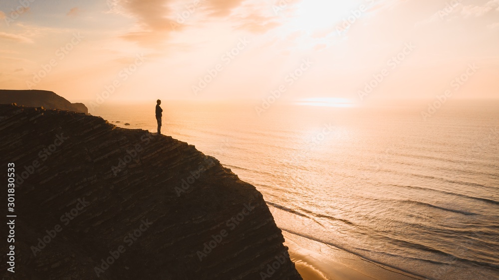 Beautiful view of a person standing on a cliff over the ocean at sunset in Algarve, Portugal - obrazy, fototapety, plakaty 