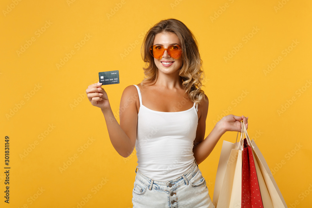 Smiling young woman girl in eyeglasses posing isolated on yellow orange background. People lifestyle concept. Mock up copy space. Holding package bag with purchases after shopping, credit bank card. Smiling young woman girl in eyeglasses posing isolated on yellow orange background. People lifestyle concept. Mock up copy space. Holding package bag with purchases after shopping, credit bank card.