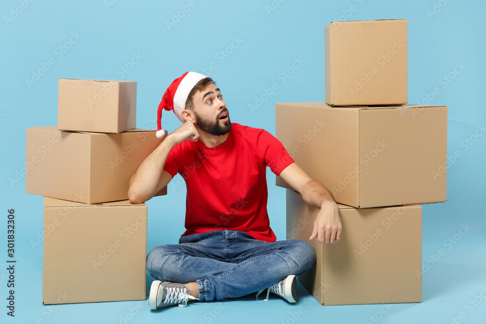 Delivery man in red uniform cap t-shirt Santa hat isolated on blue background studio portrait. Male employee courier sit cardboard box. Service concept. Mock up copy space. Happy New Year 2020 holiday Delivery man in red uniform cap t-shirt Santa hat isolated on blue background studio portrait. Male employee courier sit cardboard box. Service concept. Mock up copy space. Happy New Year 2020 holiday
